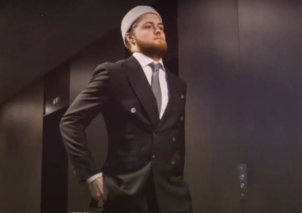 A man in a dark double-breasted suit and a white kufi cap stands confidently in an elevator, hand on hip, looking to the side.