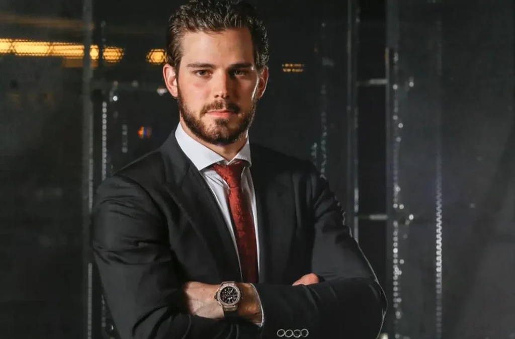 A man in a dark suit, red tie, and watch stands with arms crossed, looking confidently at the camera, set against a blurred industrial background.