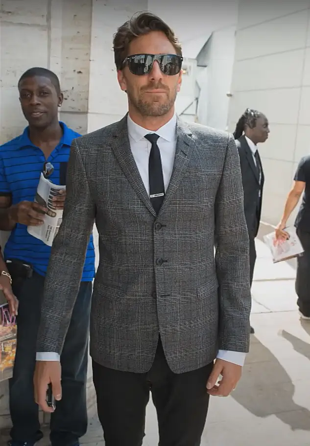 A man in a tailored grey suit and sunglasses walks confidently, with a serious expression on his face.